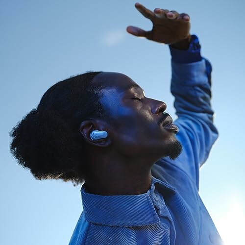 Man wearing an earbud, raising his hand against a clear sky.